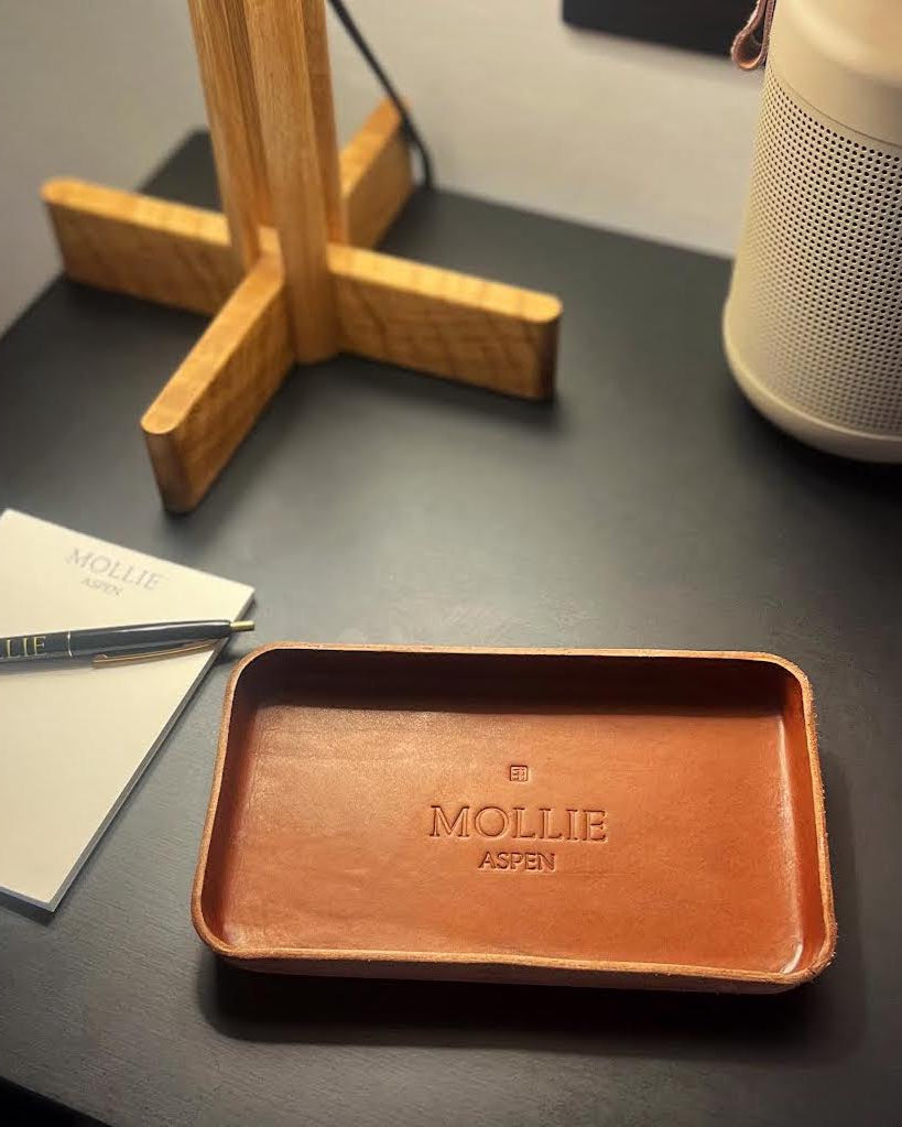 Brown tray with 'MOLLY ASPEN' branding on a dark surface with a notebook and pen.