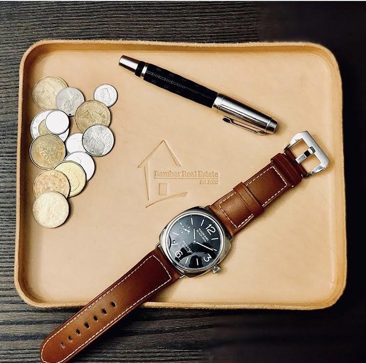Brown leather watch with a pen and coins on a beige tray with a brand logo.