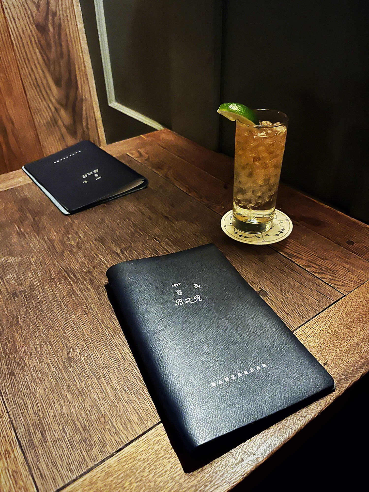Black leather notebook and a cocktail with a lime wedge on a wooden table.