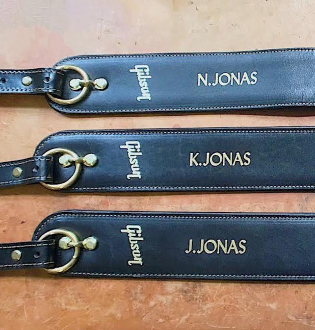 Three blue guitar straps with engraved names on a wooden surface