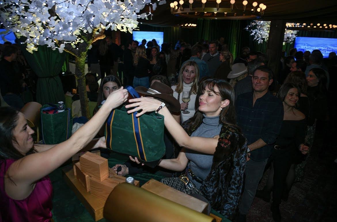 Two women interacting with a green bag in a festive indoor setting with people and decorations.
