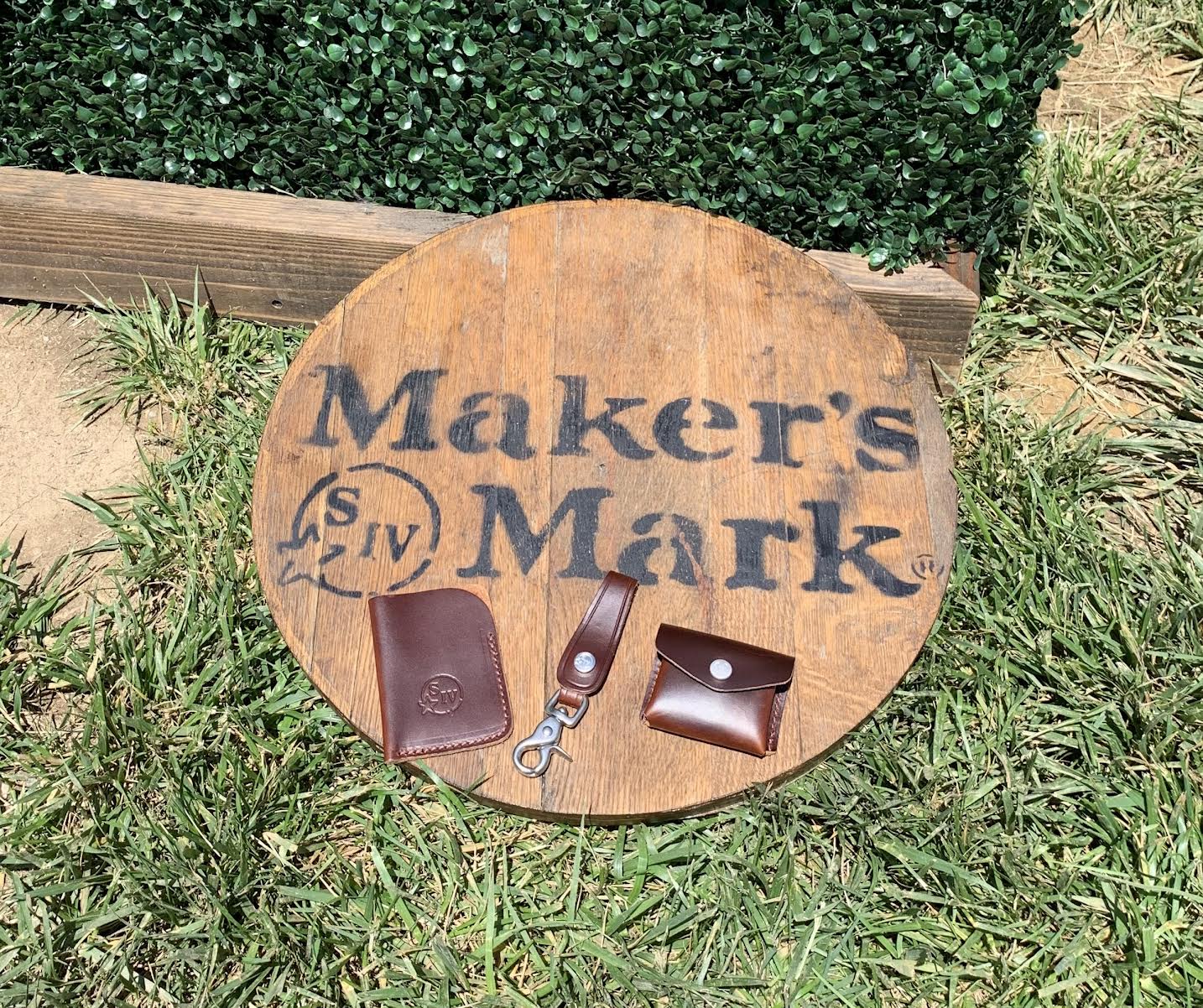 Round wooden sign with 'Maker's Mark' branding on grass; leather accessories on top