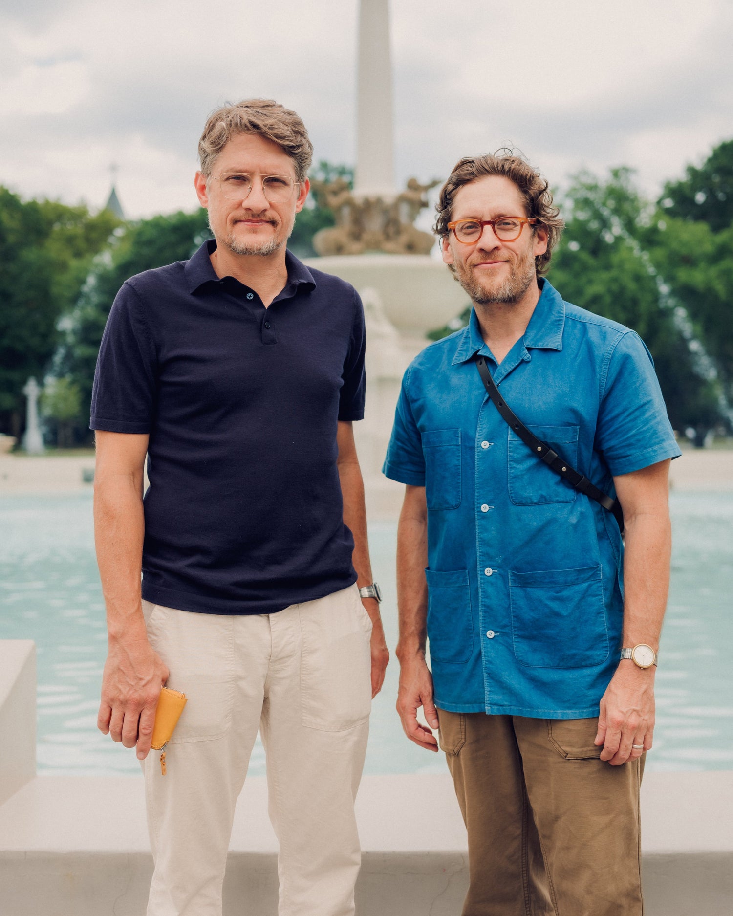 Two men standing in front of a fountain with trees and a building in the background
